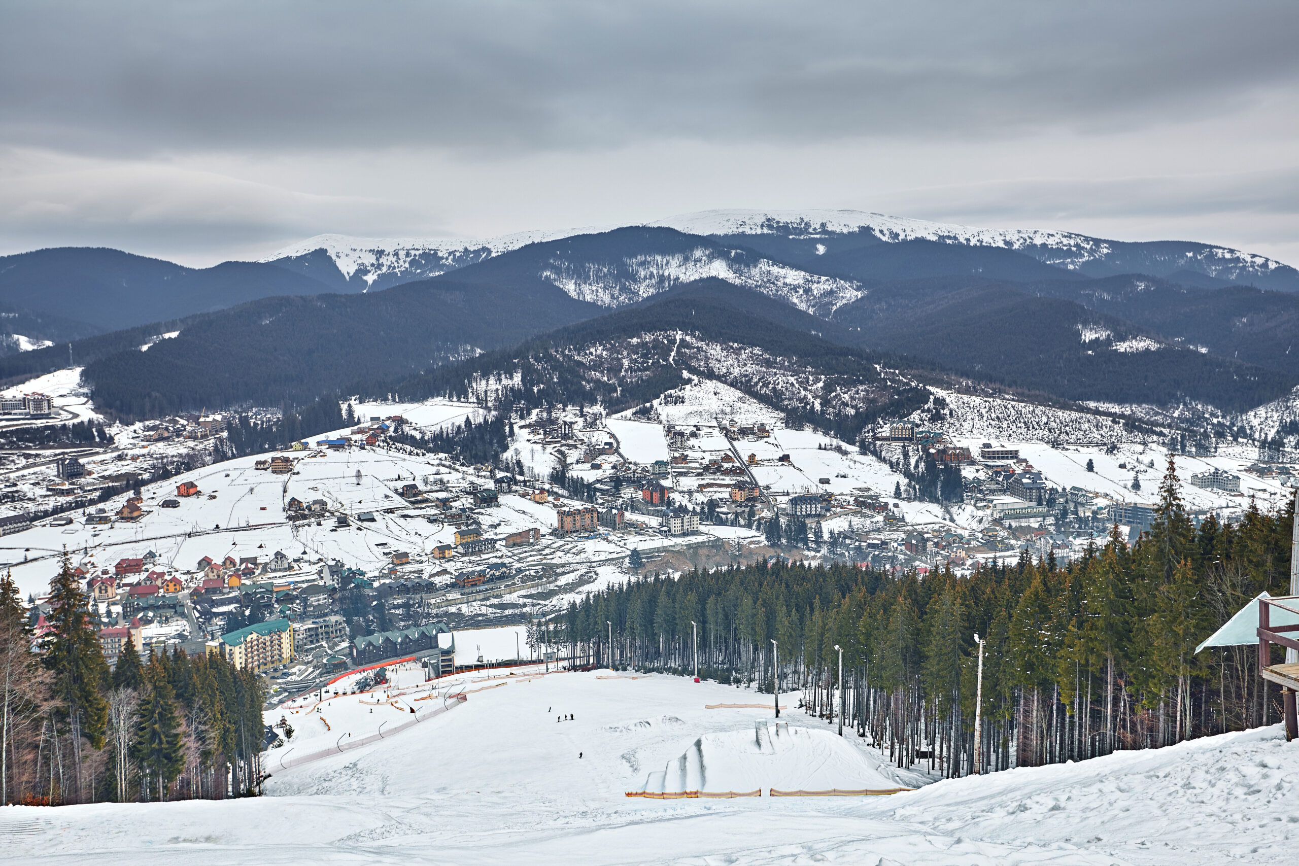 Panorama of ski resort, slope, people on the ski lift, skiers on the piste among green pine trees and snow lances.